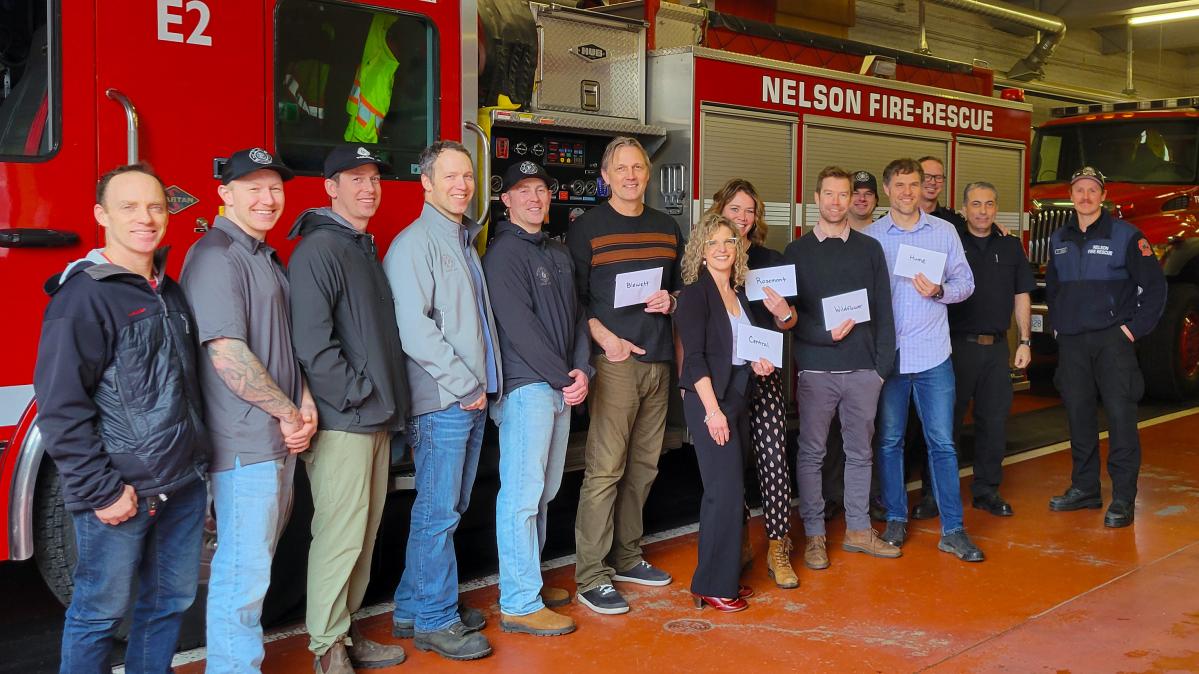 A group of people lined up in front of a fire truck inside the bay of a fire department smiling, with some holding envelopes and some in fire fighter uniforms.