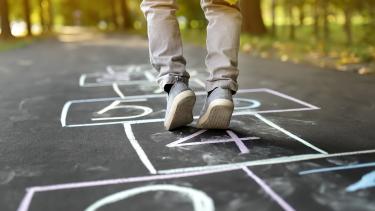 Looking at ground level at a white chalk hopscotch pattern on black paved path in a forest with only the lower legs and bottom of a yellow raincoat showing of a child stepping on a hopscotch square..