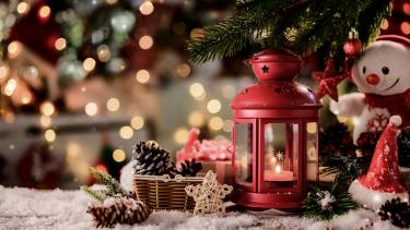 a snowman, red lantern, and pinecones on a snowy schene woth evergreen and lights in the background